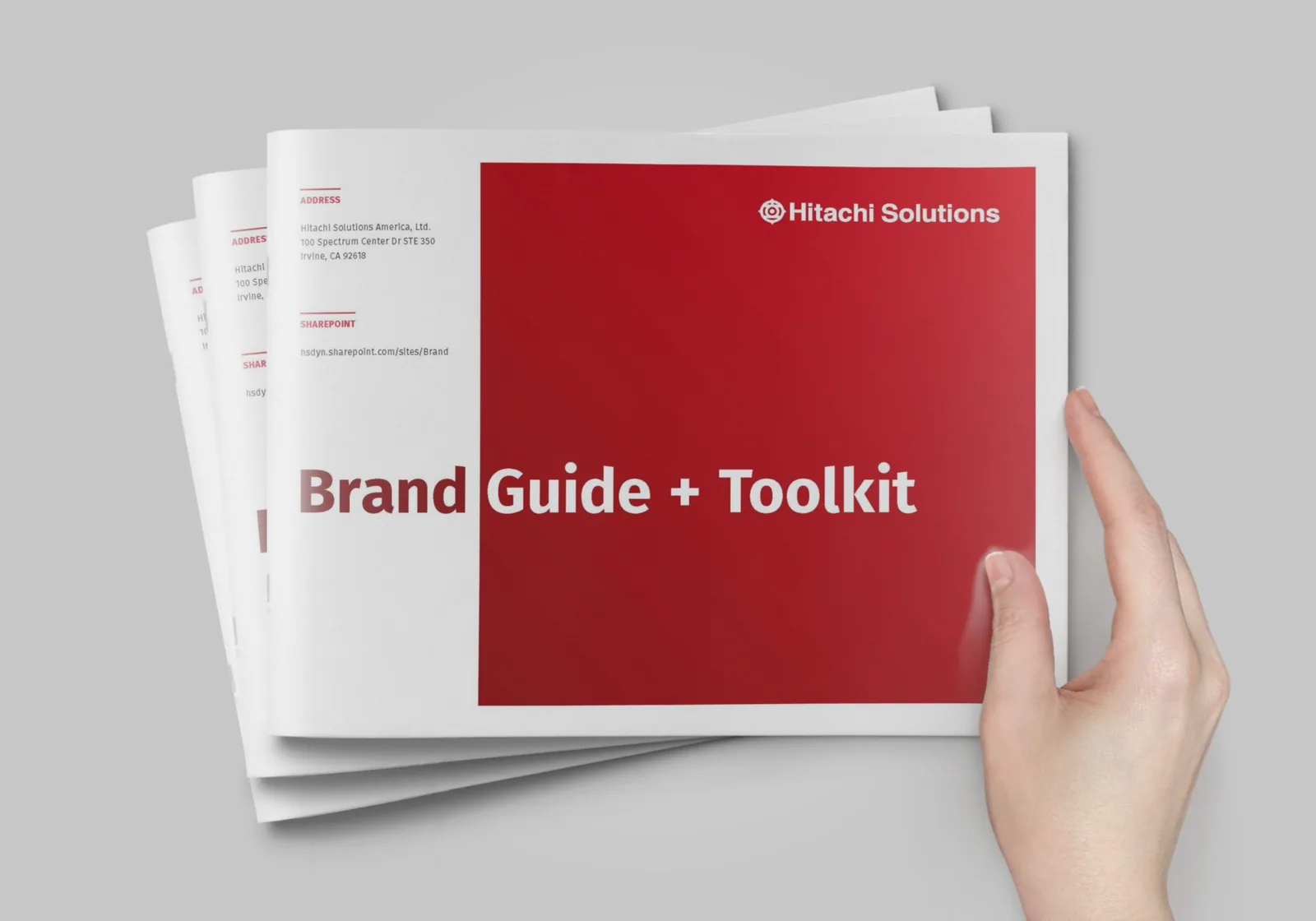 A hand holding the Hitachi Solutions Brand Guide + Toolkit, its cover in red